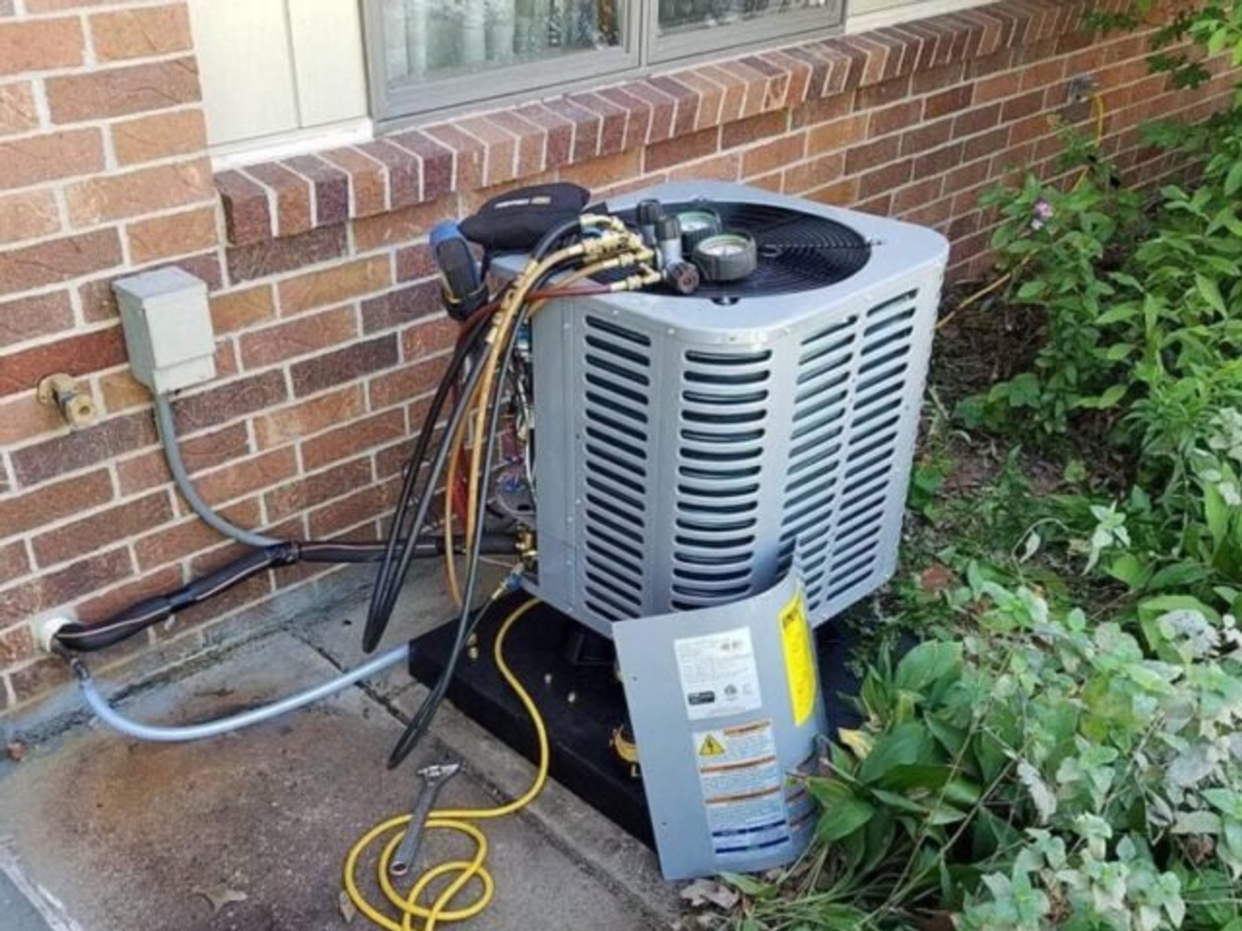 A residential condenser unit mounted on a brick wall with refrigerant lines and a service disconnect.