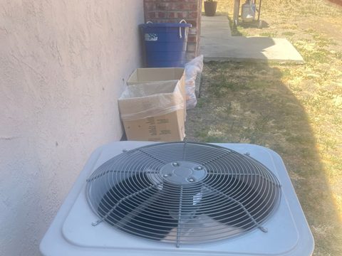 a new outdoor condenser unit sitting on the ground beside a stucco wall, with cardboard packaging and a blue recycling bin behind it